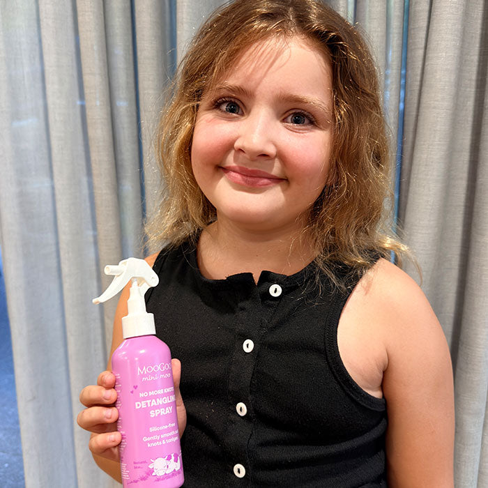 A young girl with light brown hair smiling while holding a pink bottle of MooGoo Mini Moo Detangling Spray.