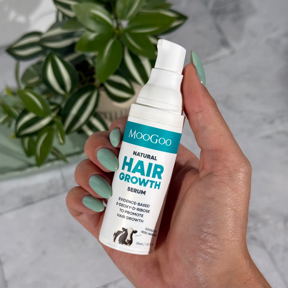 A hand holding a white pump bottle of MooGoo Natural Hair Growth Serum, with a green potted plant in the background on a light marble surface.