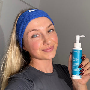 A blonde woman wearing a blue terry towelling Headband with a MooGoo label. The woman is also holding the MooGoo Oil Cleanser in her hand.