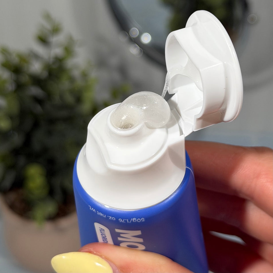 Hand holding MooGoo Mini Moo Kids Blueberry Smoothie flavoured toothpaste, with a toothbrush greenery and blue tiles in the background. Healthy habit building, tastes like blueberries. Fluoride-free formula, safe for kids. Safe if Swallowed. 