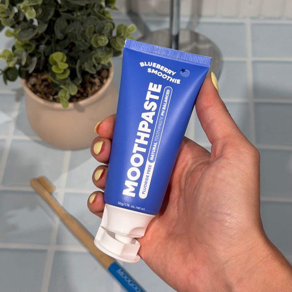 Hand holding a tube of MooGoo Mini Moo Blueberry Smoothie kids toothpaste and squeezing out the clear toothpaste. Healthy habit building, tastes like blueberries. Fluoride-free formula, safe for kids. Safe if swallowed. 