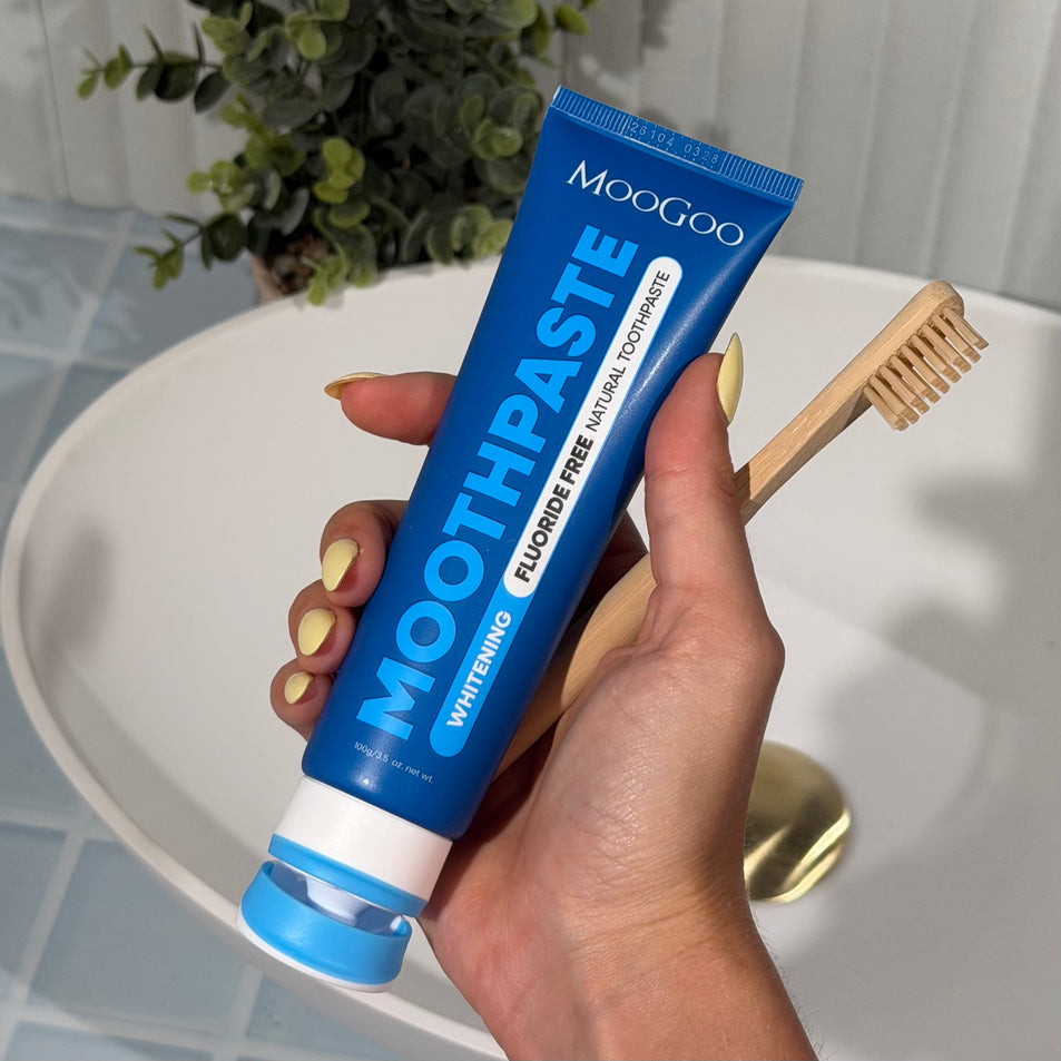 Hand holding a tube of MooGoo Fluoride Free Peppermint Whitening toothpaste with a wooden toothbrush in a bathroom setting.