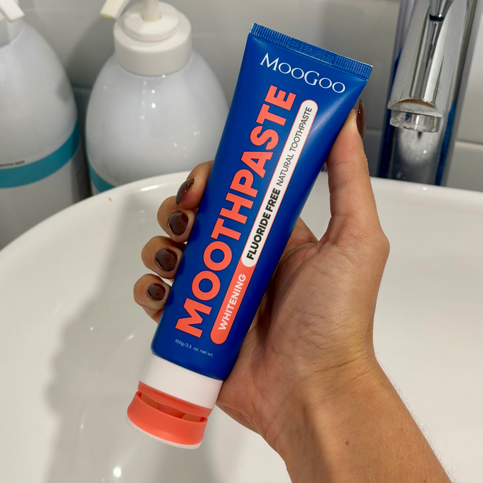 Hand holding a tube of MooGoo Fluoride Free Watermelon and Mint Peppermint Whitening toothpaste h in a bathroom setting.